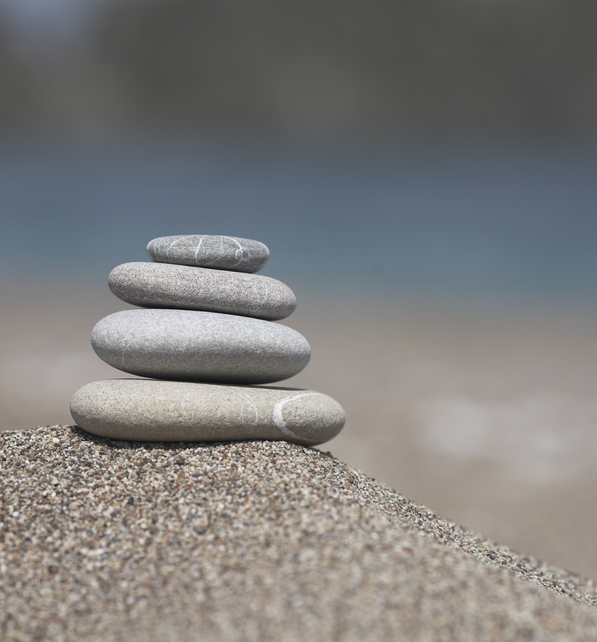 Stack of five smooth, rounded stones of varying sizes and colors, arranged from smallest on top to largest at the bottom, placed on a bed of multicolored sand with a blurred grayish background.