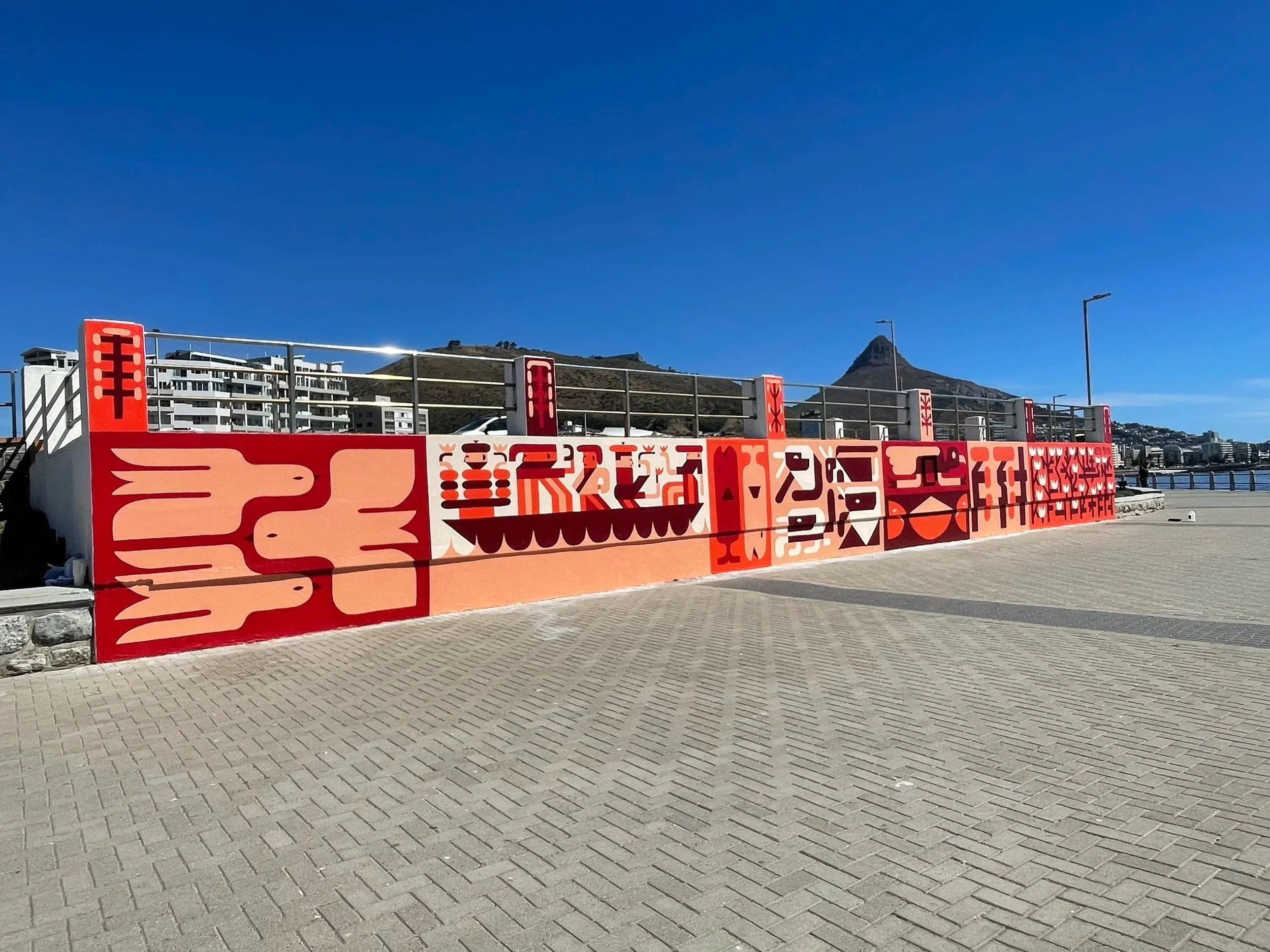 Colorful mural with red and pink African-inspired patterns on a concrete wall in an outdoor area with a mountain and blue sky in the background.