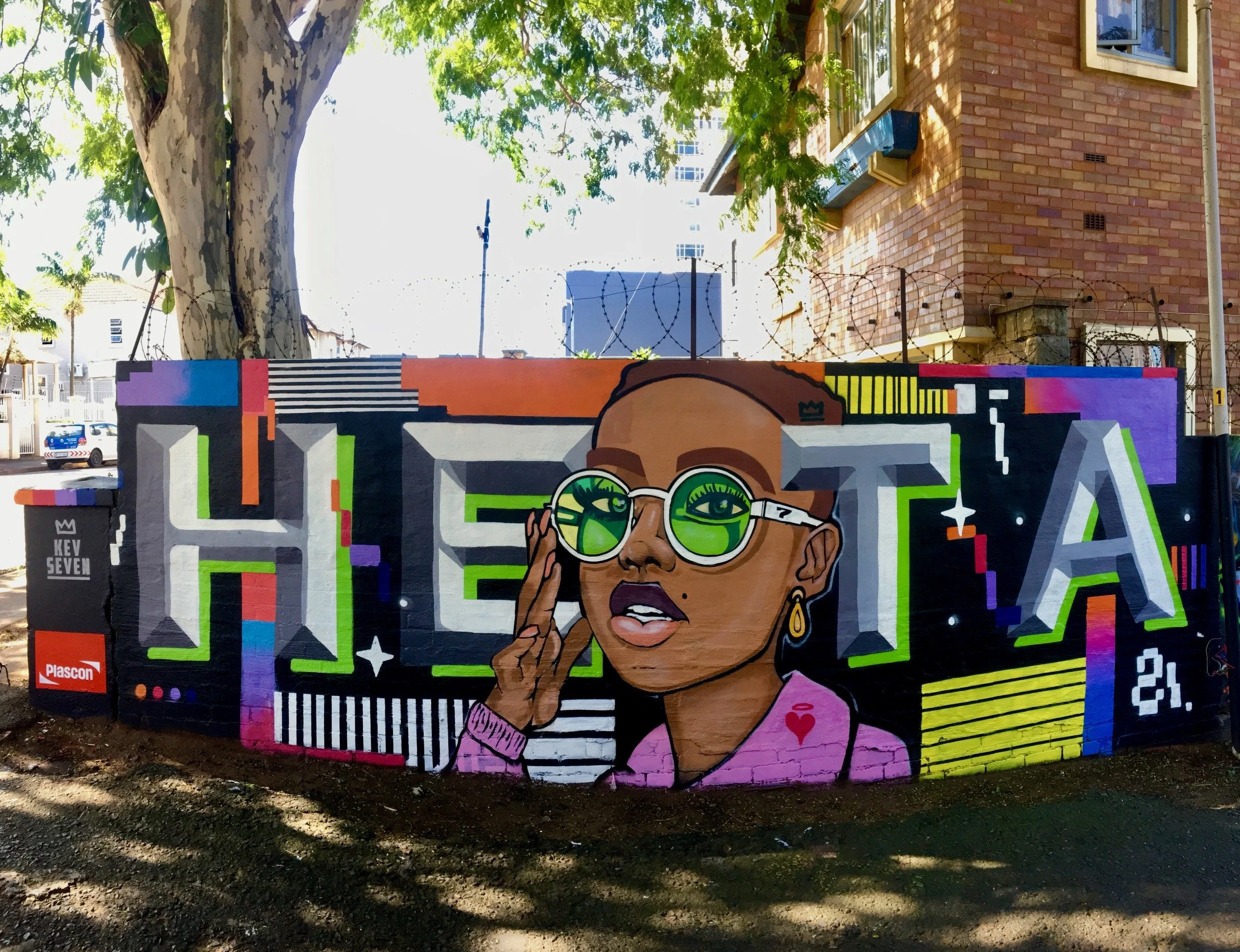 Colorful mural graffiti artwork of a woman with glasses, holding her hand near her face, on a brick wall in an urban setting with trees and buildings in the background.