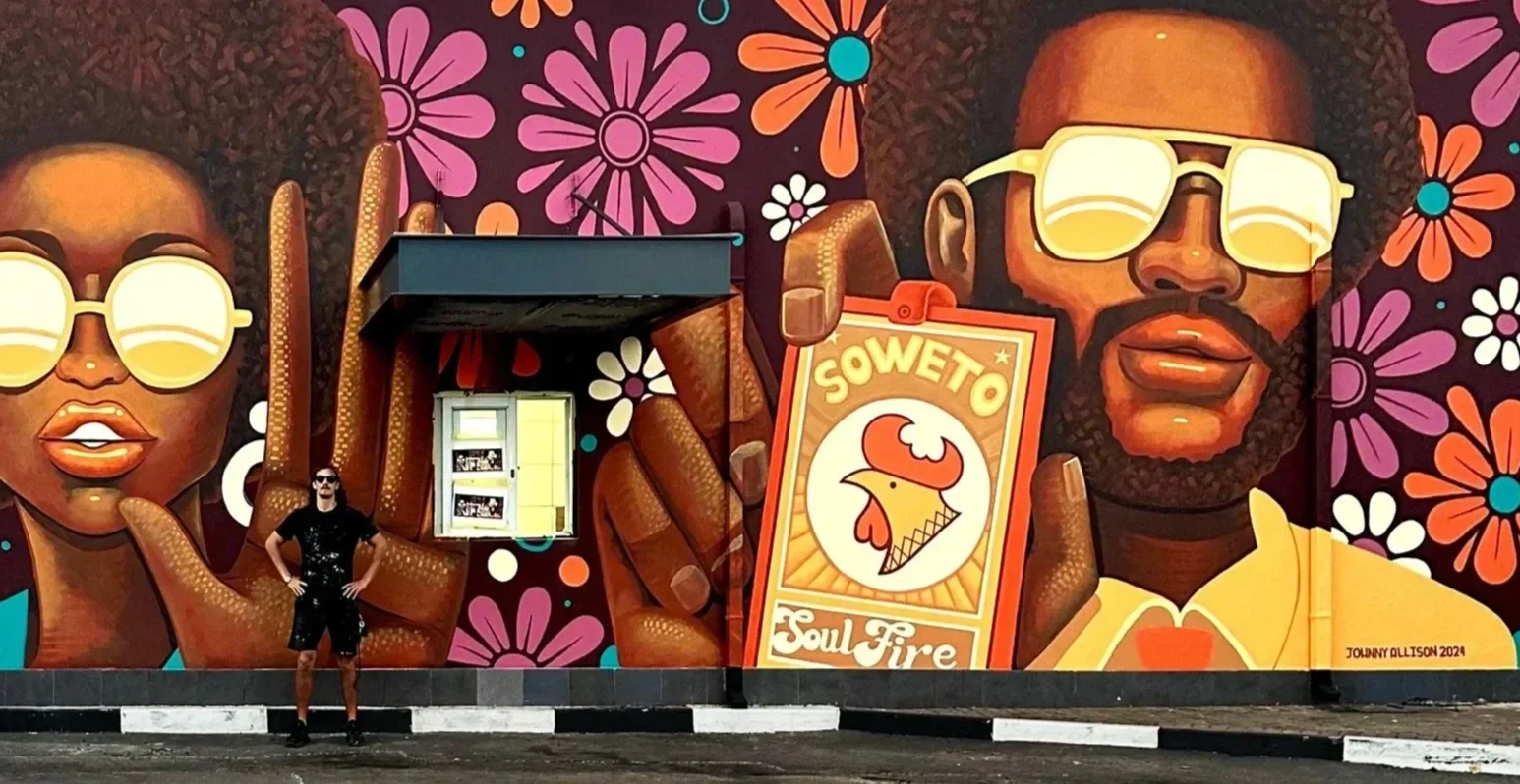 A person in black clothes and sunglasses standing in front of a large colorful mural depicting two African American individuals with large sunglasses, surrounded by vibrant flowers, and holding a sign that says "Soweto Soul Fire".