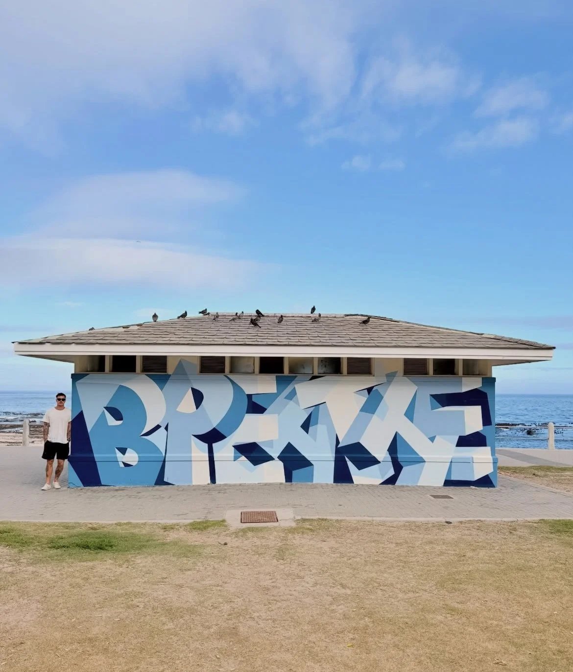 A man standing next to a building with a large blue and white graffiti mural, located near the beach with rocks and the ocean in the background. There are seagulls on the roof of the building and a clear sky above.