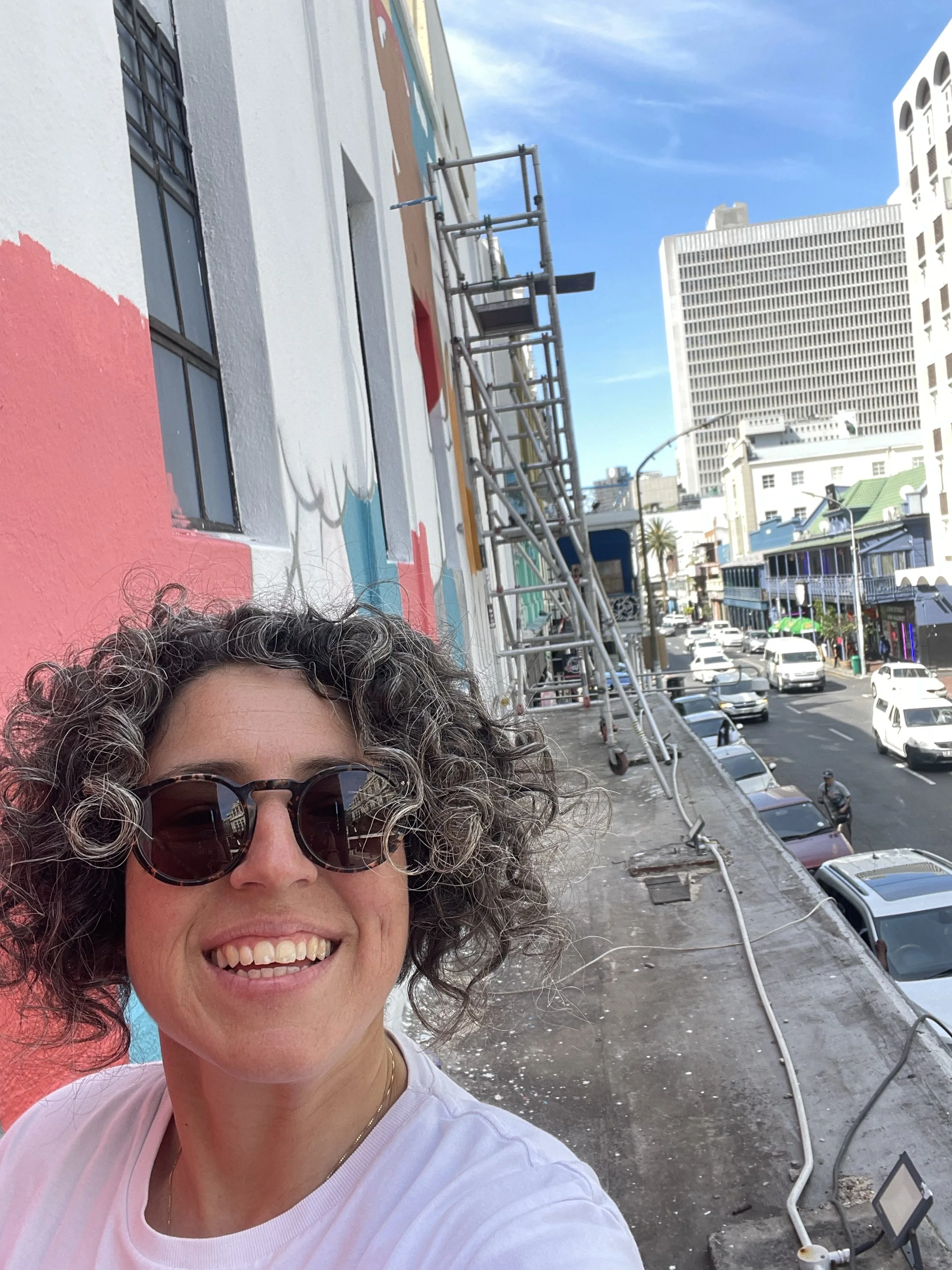 A woman with curly hair and sunglasses takes a selfie on the street near a wall with colorful mural. The mural is being painted and scaffolding is visible. Cityscape with tall buildings and vehicles is in the background.