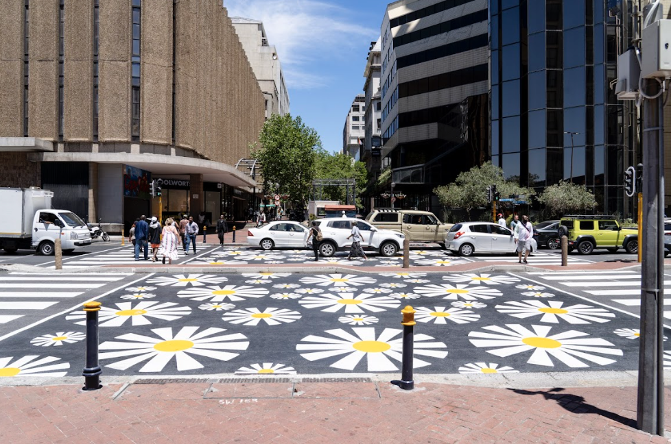 City street crosswalk with large white and yellow daisy flower pattern, surrounded by cars and pedestrians, modern buildings on both sides, sunny day.