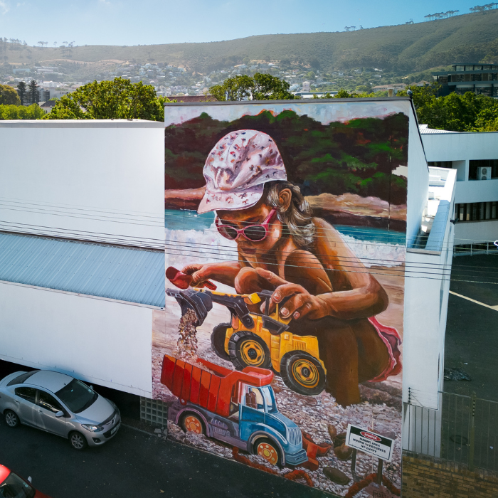 Large mural of a girl playing with toy dump trucks at the beach, painted on the side of a building.