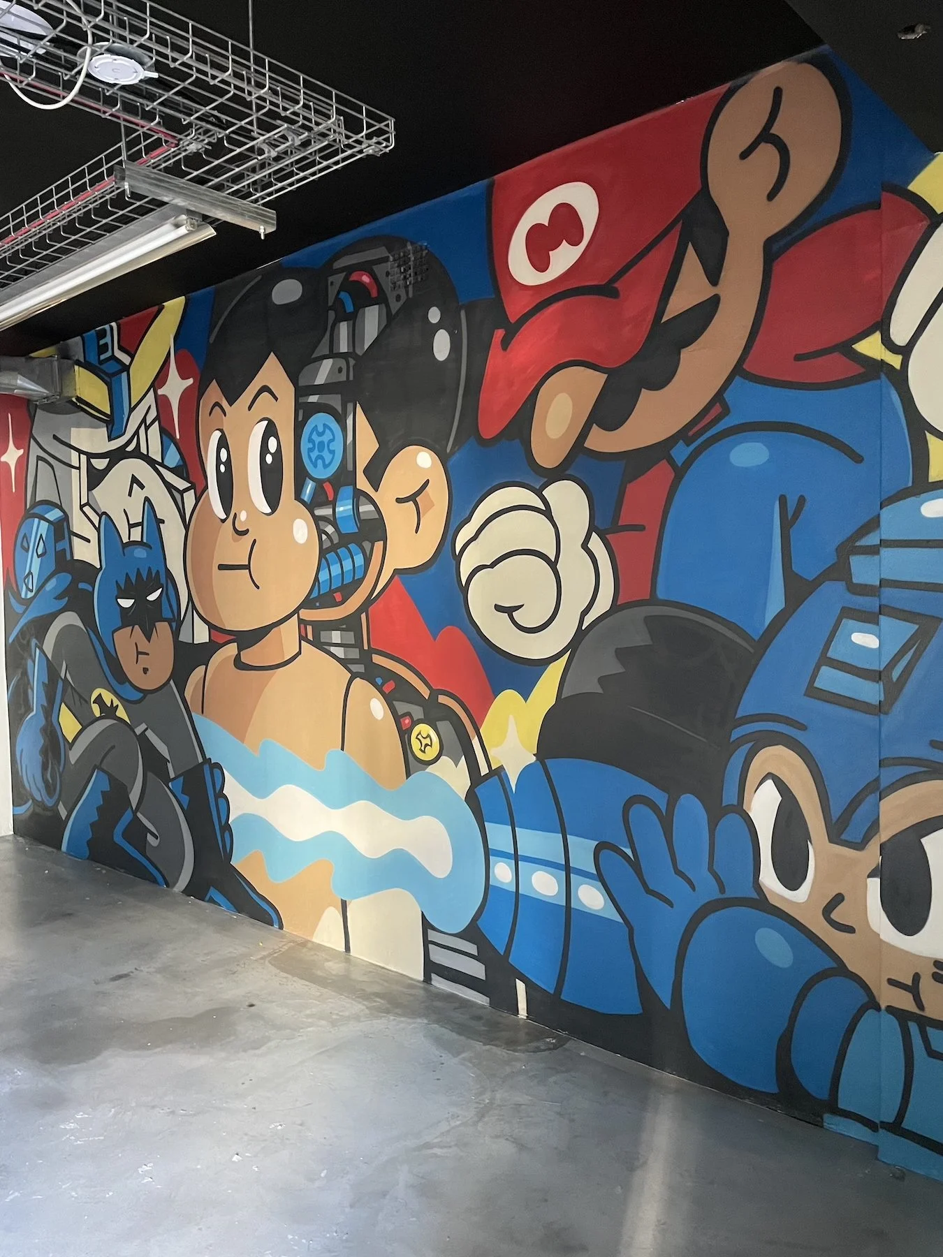 Colorful graffiti mural featuring cartoon characters, including a girl with headphones, a superhero, and a large character resembling Mario from Nintendo.