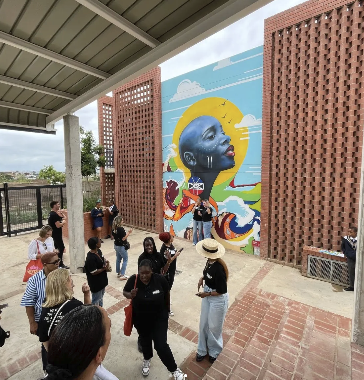 A group of diverse people gathered in an outdoor area near a colorful mural of a woman with dark skin, short hair, and hoop earrings. The mural features vibrant colors, clouds, a yellow sun, and a landscape.