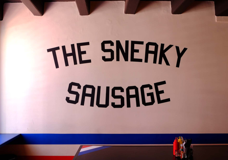 Wall display with the words 'The Sneaky Sausage' in black, uppercase letters across a pink wall, with condiments and utensils on a counter below.