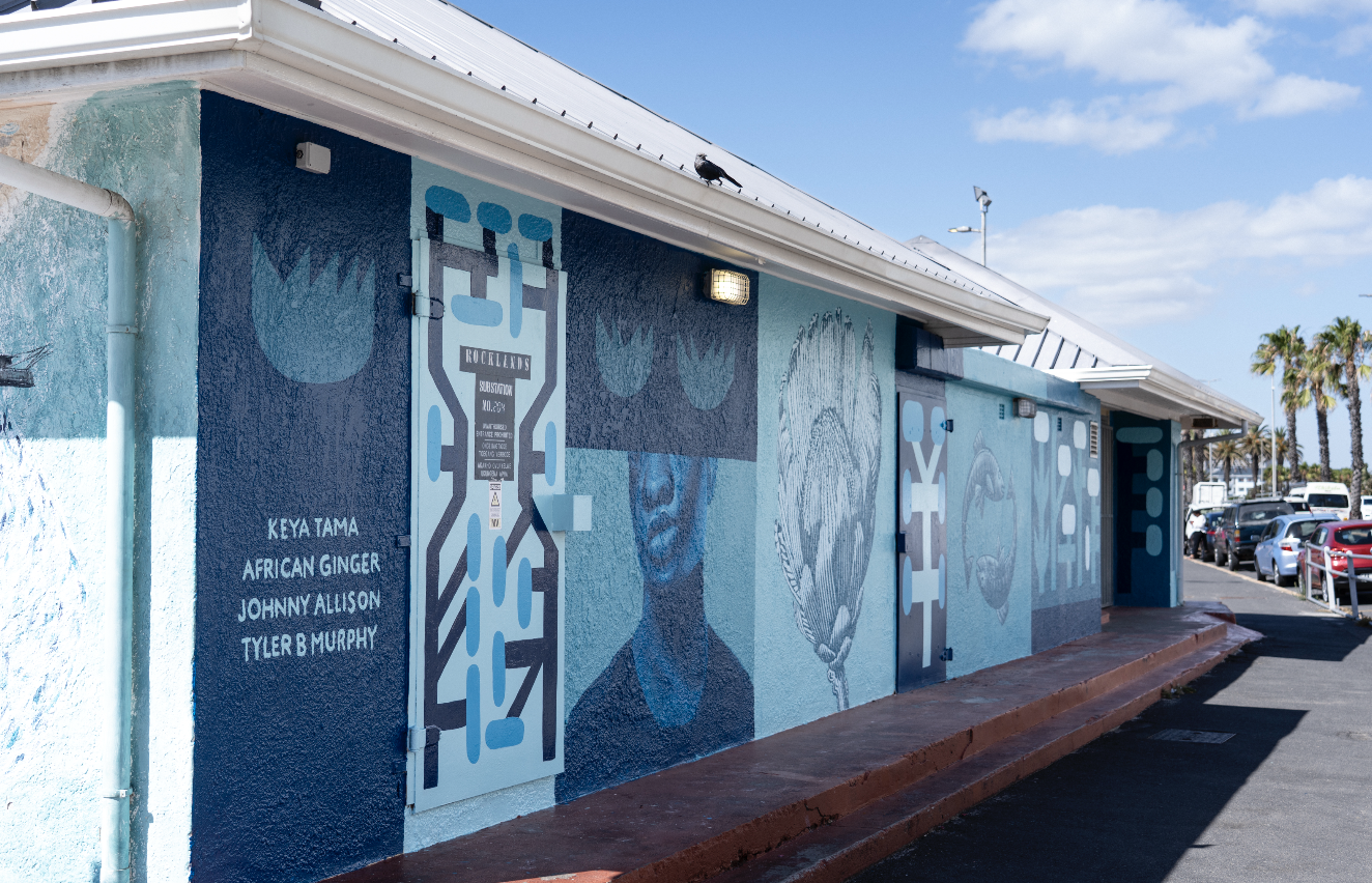 Colorful mural on the side of a building featuring abstract faces, symbols, and names of artists, with a row of parked cars and palm trees in the background on a sunny day.