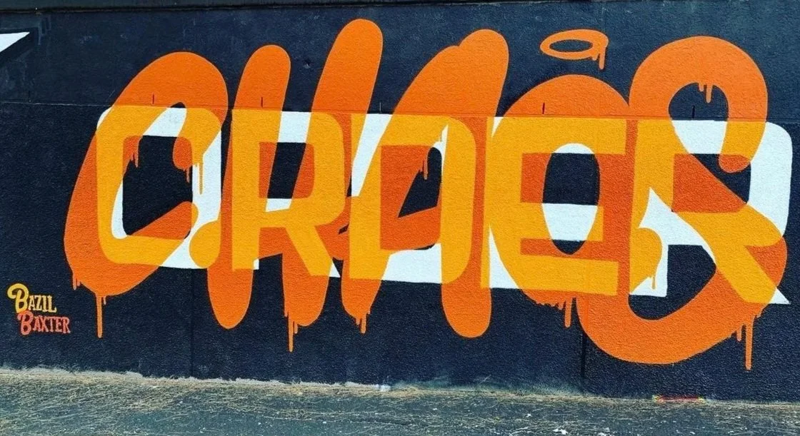 Orange graffiti spelling 'CRUES' on a dark wall with smaller artist signatures 'Bazzl' and 'Baxter' in the bottom left corner.