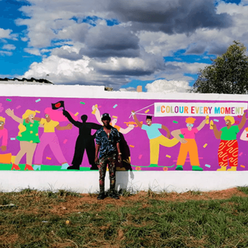 A person standing in front of a colorful mural with various animated characters and the text 'COLOUR EVERY MOMENT' written on it, under a partly cloudy sky.