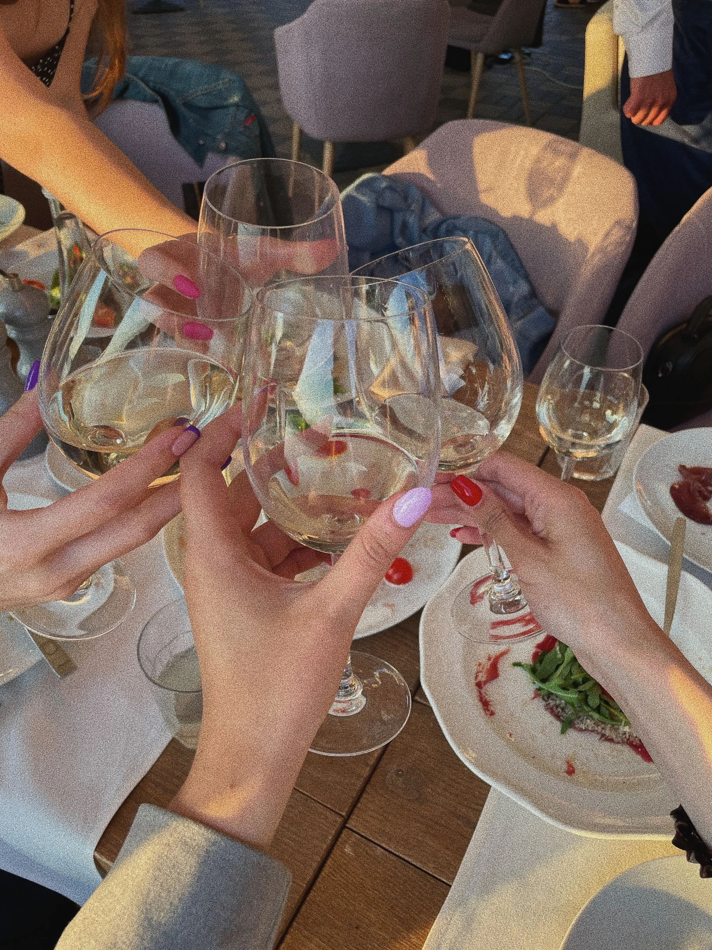 Several glasses of white wine being clinked together in a toast at a dining table with plates of food in the background.