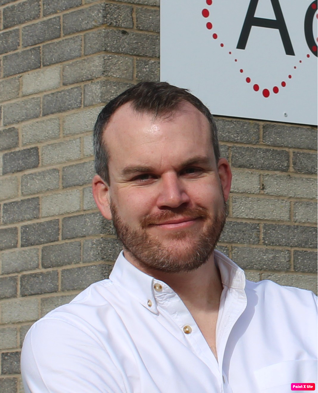 A smiling man with short brown hair and a beard, wearing a white collared shirt, standing outdoors against a brick wall with a partial sign visible in the background.
