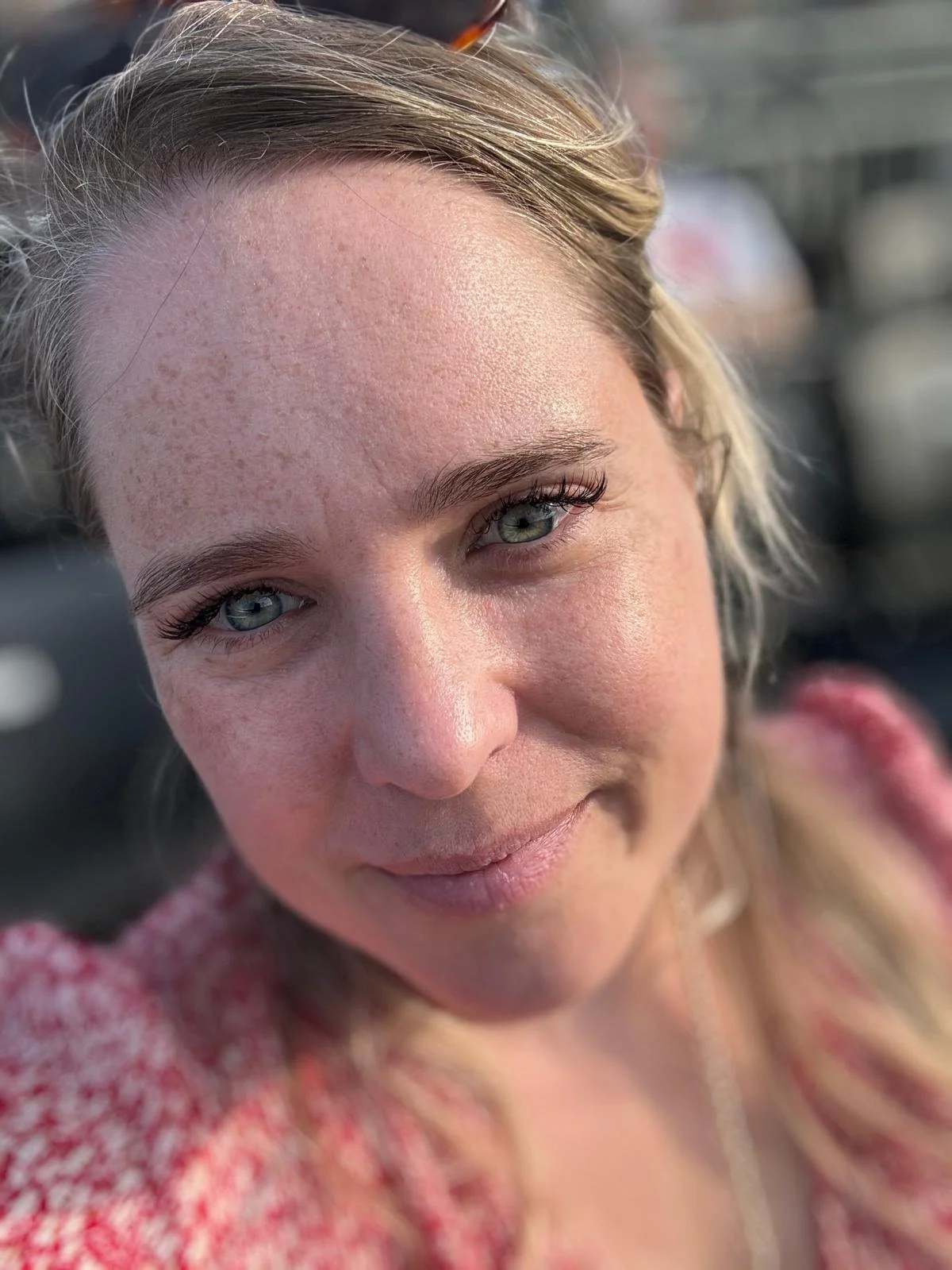 Close-up van een lachende vrouw met blauwe ogen en blonde haar, gekleed in een roze en witte kleding, buiten op een zonnige dag.