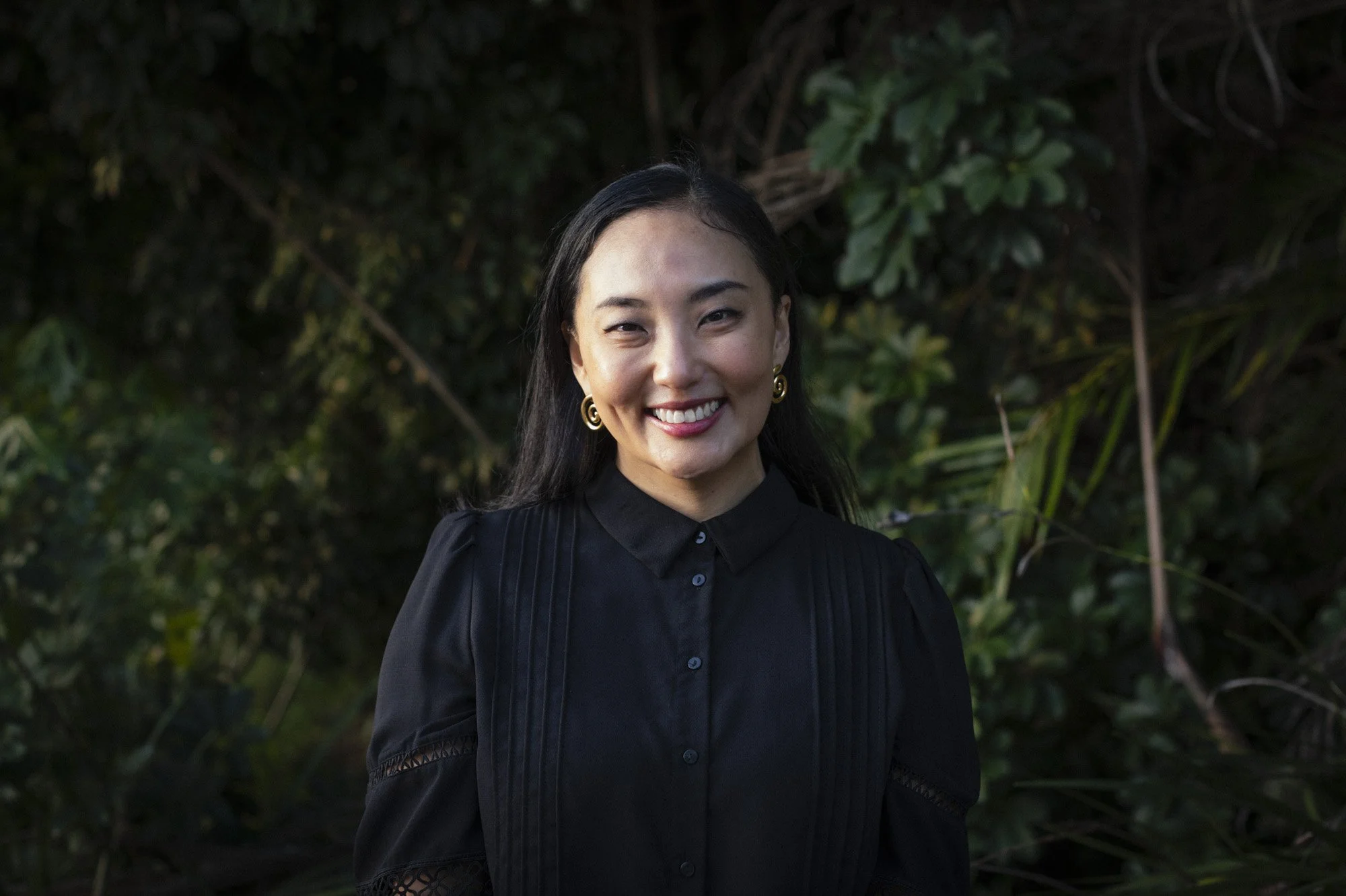 A woman with long dark hair, smiling, wearing gold earrings and a black shirt with lace details on the sleeves, standing outdoors in front of a lush green leafy background.