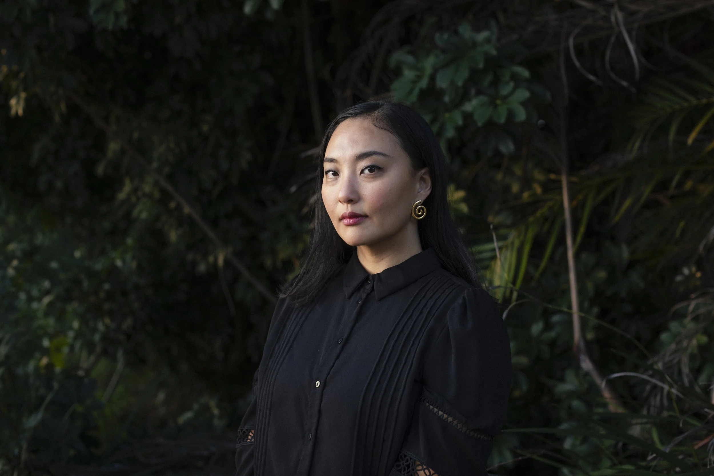 A woman with long black hair, wearing a black shirt and large spiral gold earrings, posing outdoors with dark green foliage in the background.