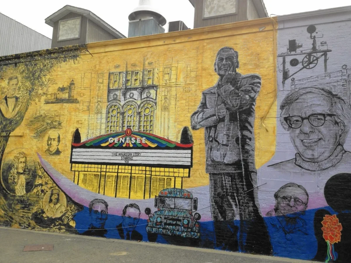 Colorful mural featuring historical figures, a movie marquee reading 'Genesee' and 'The Waukegan Story Starring You,' along with an old vehicle, and artistic sketches of buildings, people, and a film camera on a brick wall.
