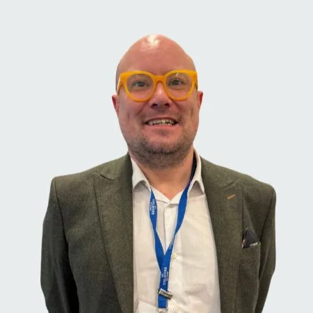 A man with a bald head and glasses smiling, wearing a blazer and a lanyard, against a plain light background.