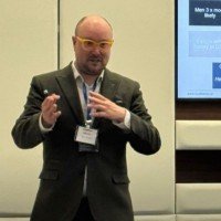 A man in a suit giving a presentation, holding a cup, with a screen displaying a slide in the background.