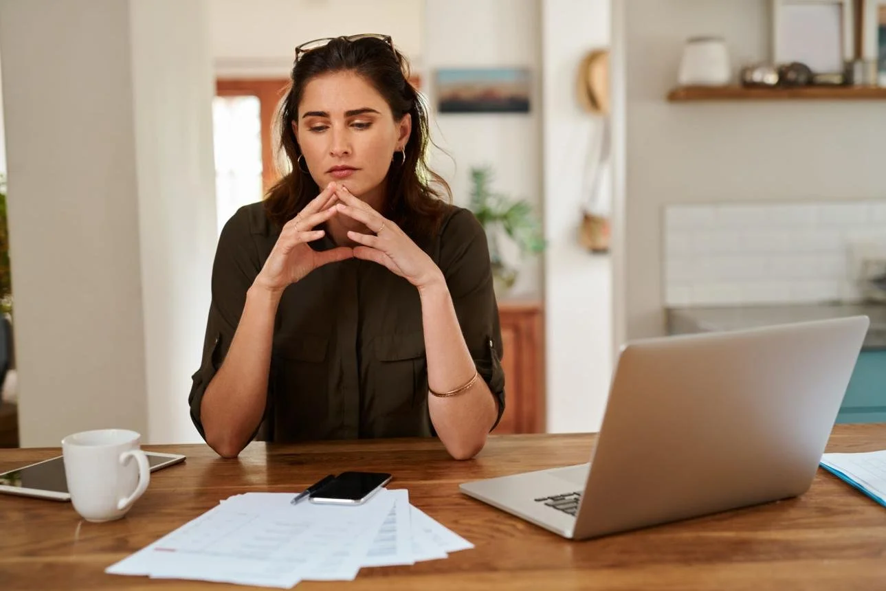 Woman sitting at a wooden table with a laptop, papers, a smartphone, a tablet, and a coffee mug in a bright kitchen.