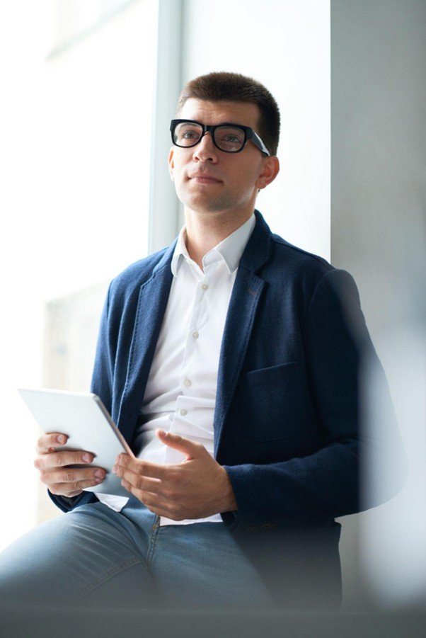 Young man with glasses in business attire sitting by a window, holding a tablet, looking confident.