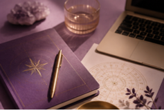 Laptop, glass of whiskey, closed purple notebook with gold star, purple crystal, and decorative paper on a desk.