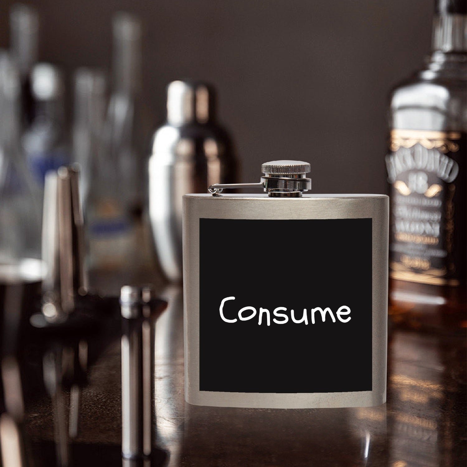A metal flask with a black label reading 'Consume' in a bar or lounge setting with various bottles and bar tools in the blurred background.