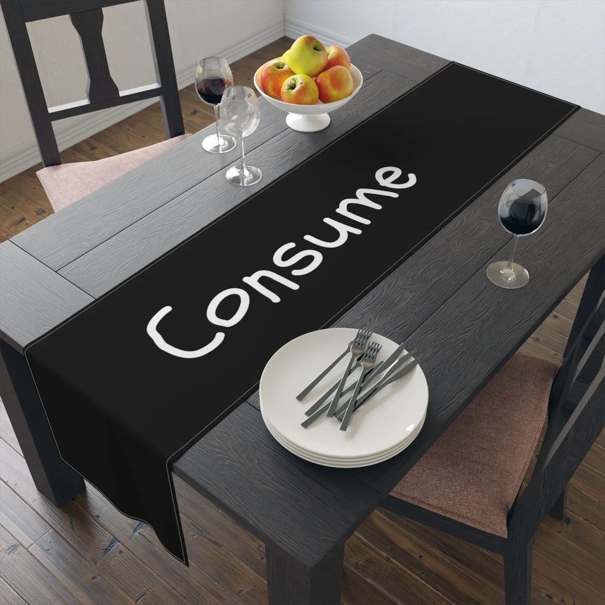Dining table with a black table runner labeled 'Consume,' a white bowl of apples, two glasses of red wine, set of white plates, and forks.