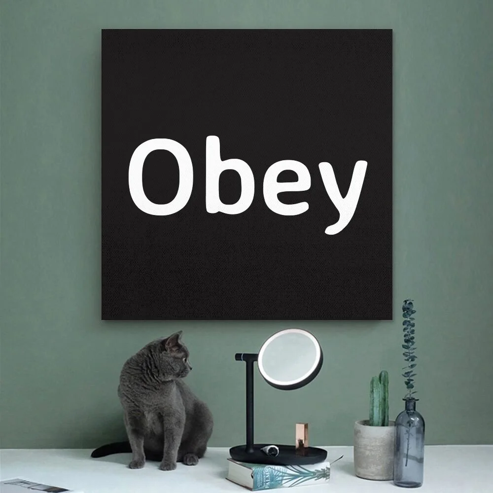 A modern room with a green wall, a black artwork with the word 'Obey' written in white, a gray cat sitting on a white desk, a round mirror with a black stand, a potted cactus, a clear gray bottle with a plant, and a hardcover book.