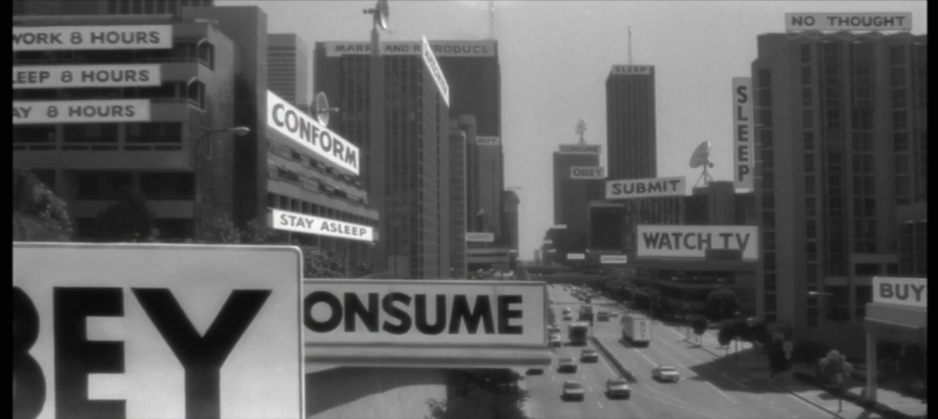 A city street with numerous billboards and signs displaying messages such as "CONFORM," "STAY ASLEEP," "SUBMIT," "OBEY," "WATCH TV," and "BUY." Cars are driving on the multi-lane road below the signs.
