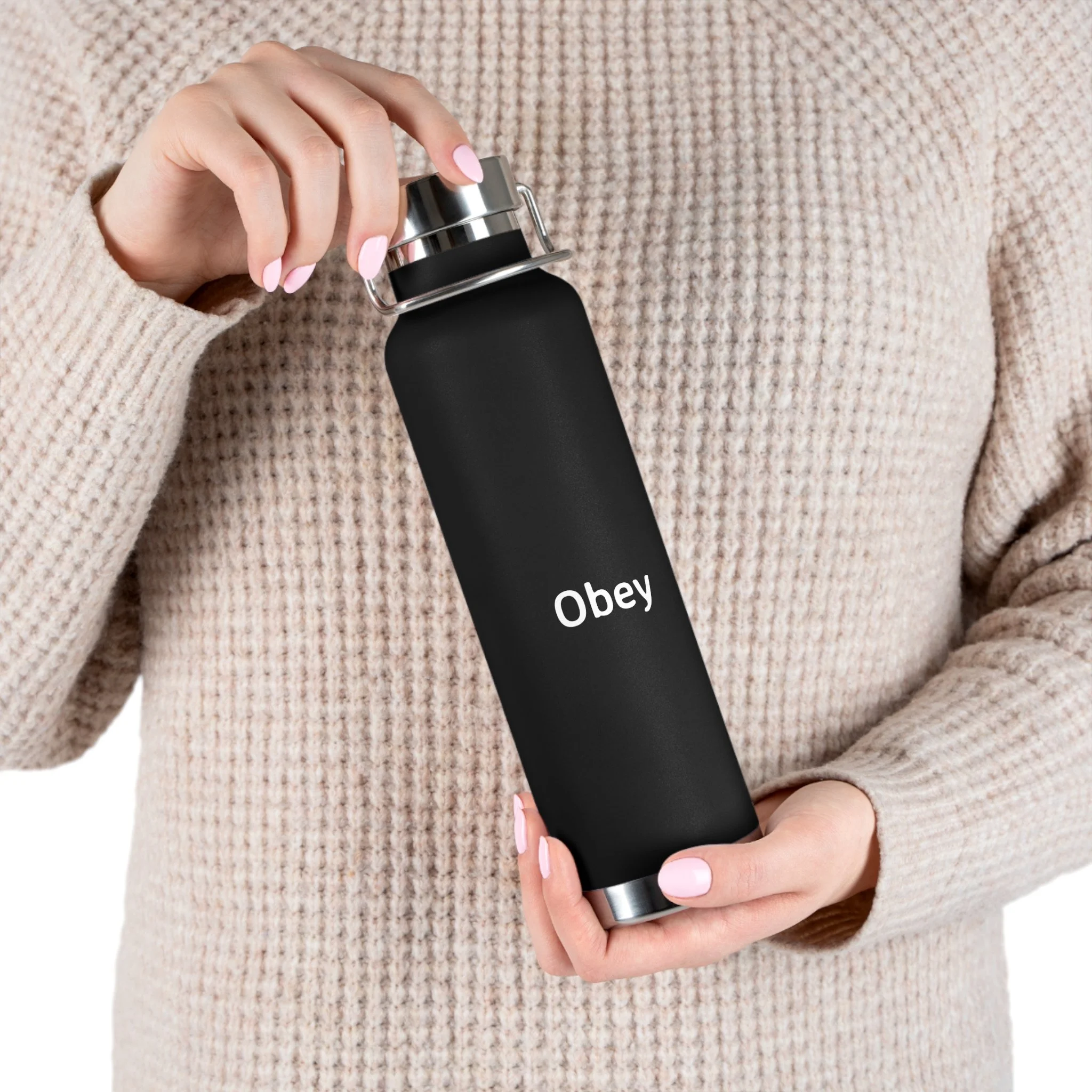 Person holding a black water bottle with the word 'Obey' printed on it, wearing a beige textured sweater.