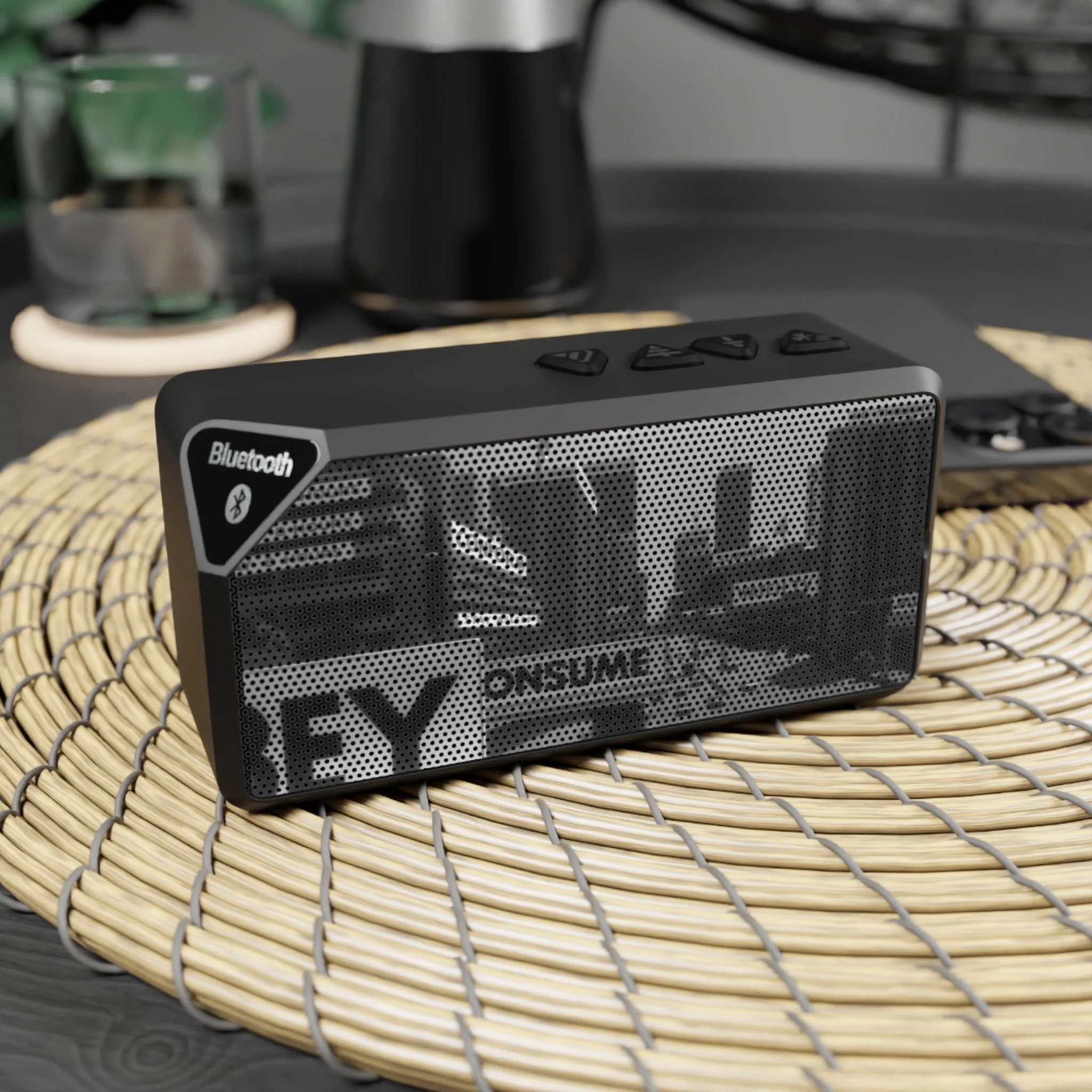 A portable Bluetooth speaker with a mesh front displaying the words "YES ONSUME" on a woven placemat on a table.