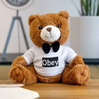 A teddy bear wearing a white T-shirt with the word 'Obey' and a black bow tie, sitting on a wooden surface.