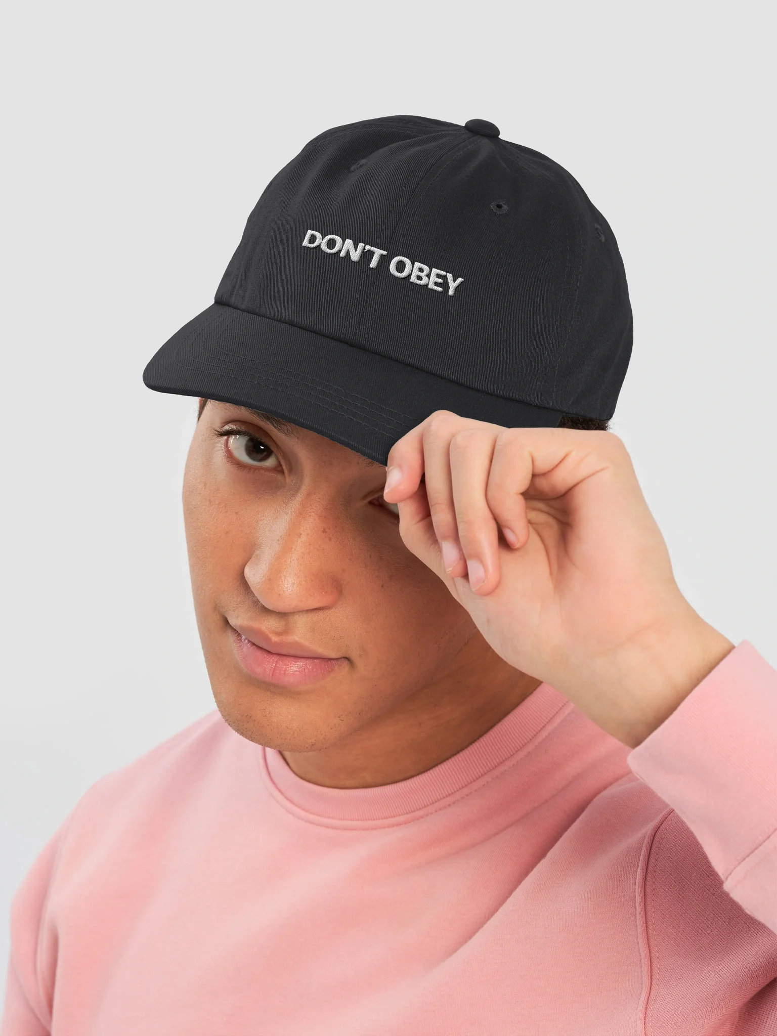 A young man wearing a pink sweatshirt and a black baseball cap that says 'DONT OBEY,' tipping the cap with his right hand against a light background.