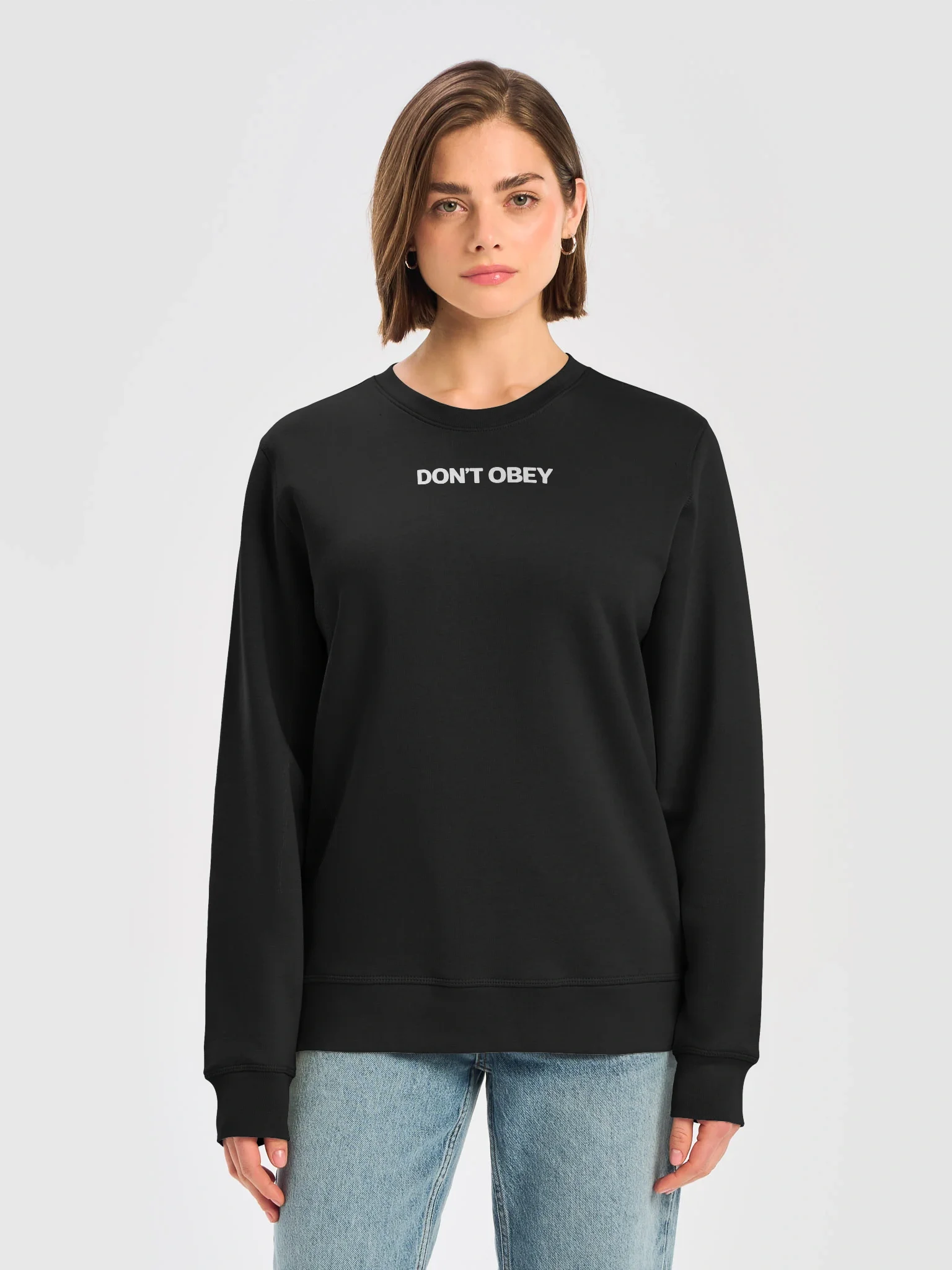 A woman with shoulder-length brown hair wearing a black sweatshirt with the words "DON'T OBEY" printed on it, and light blue jeans.