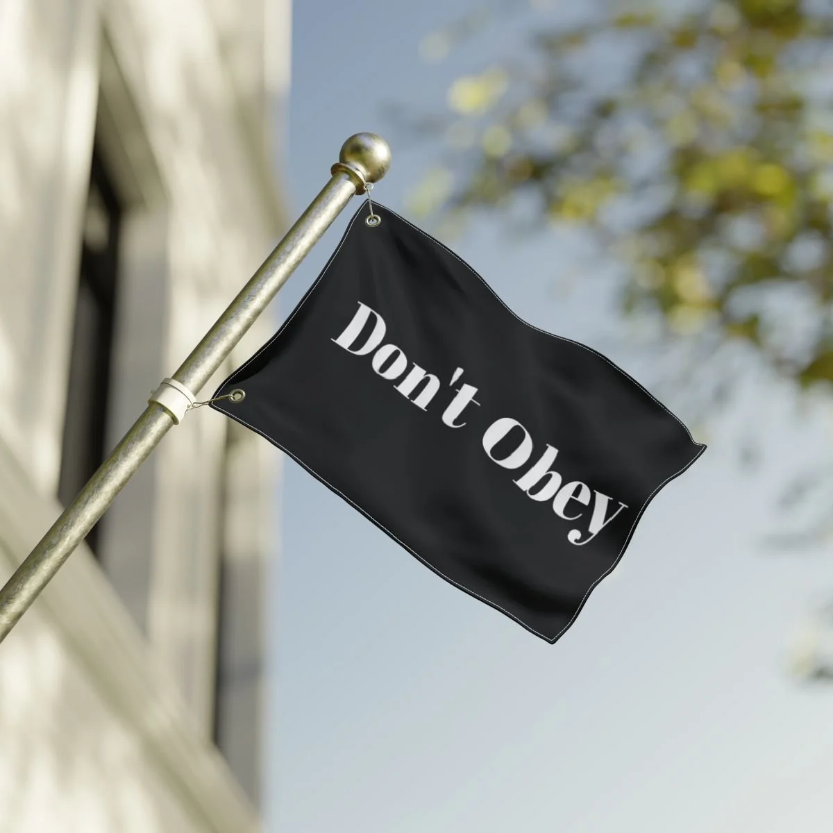 A black flag with white text that says "Don't Obey" is hanging from a metal pole against a background of a building and tree with leaves.