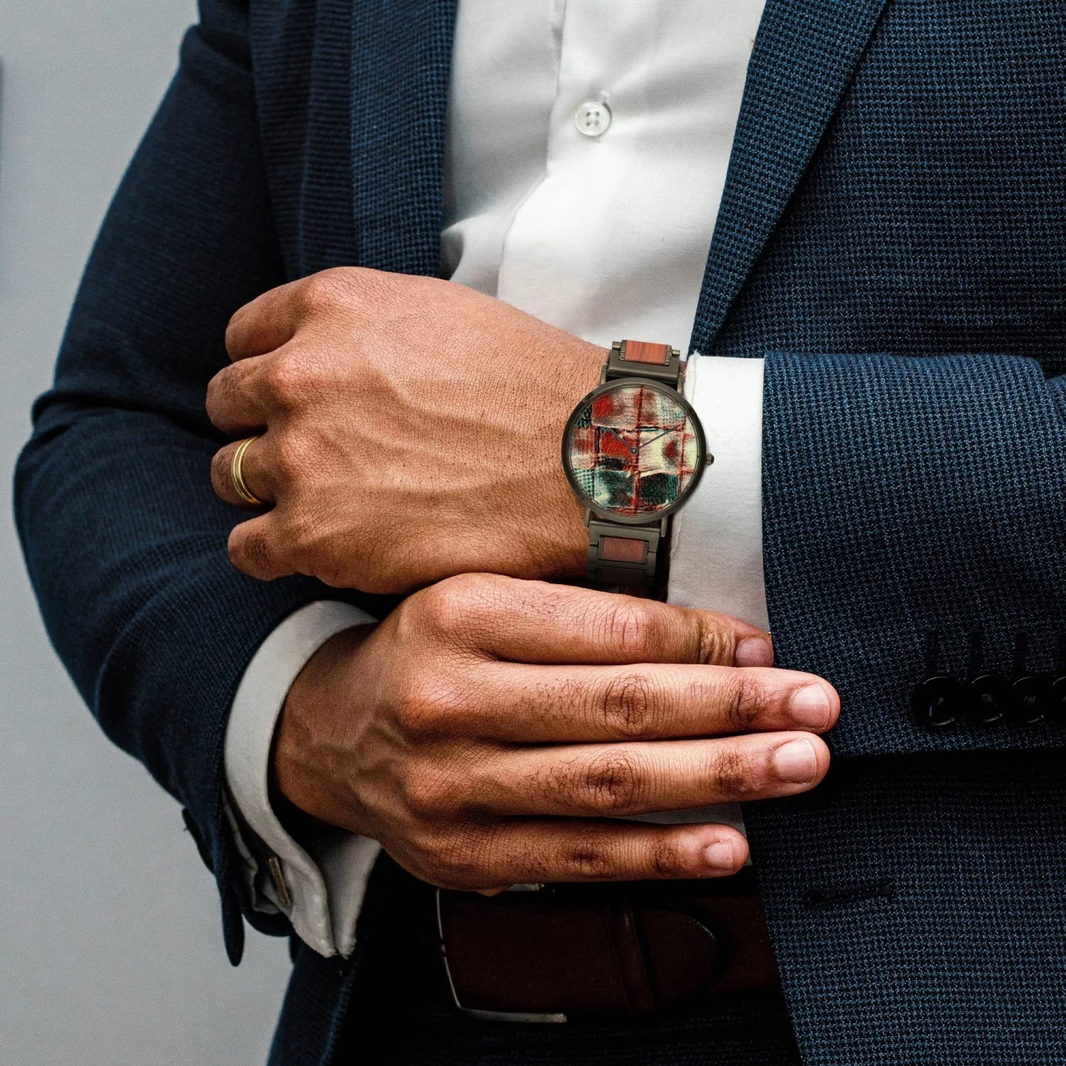 A man in a dark blue suit with a white shirt, wearing a wristwatch with a colorful abstract design, and a gold wedding band on his left ring finger. His right hand rests over his left wrist, displaying the watch.