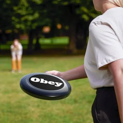 Person holding a round black frisbee with the word 'Obey' printed on it in white, outdoors in a park.