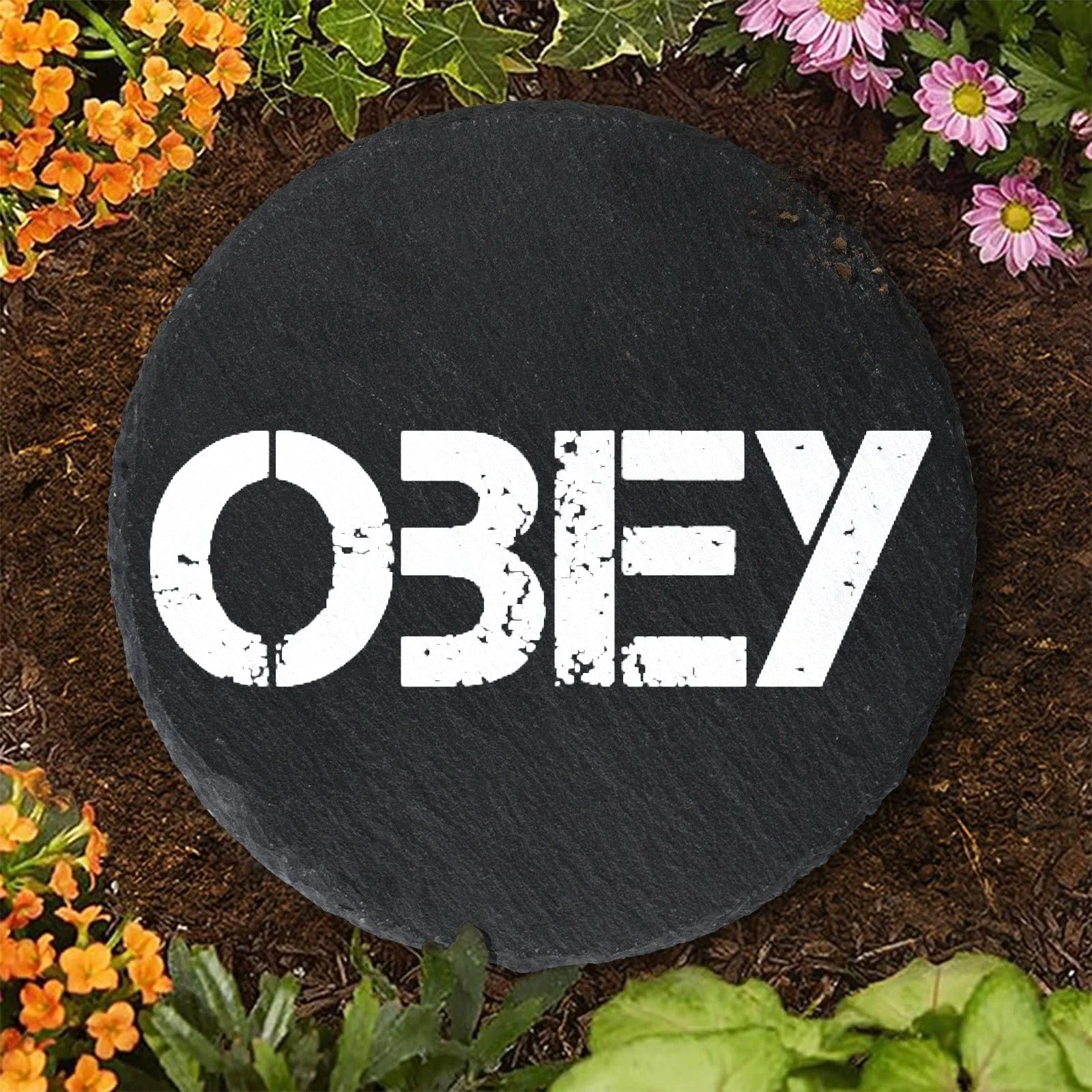 A round black sign with the word 'OBEY' in white, distressed block letters, placed on the soil of a garden bed surrounded by colorful flowers.