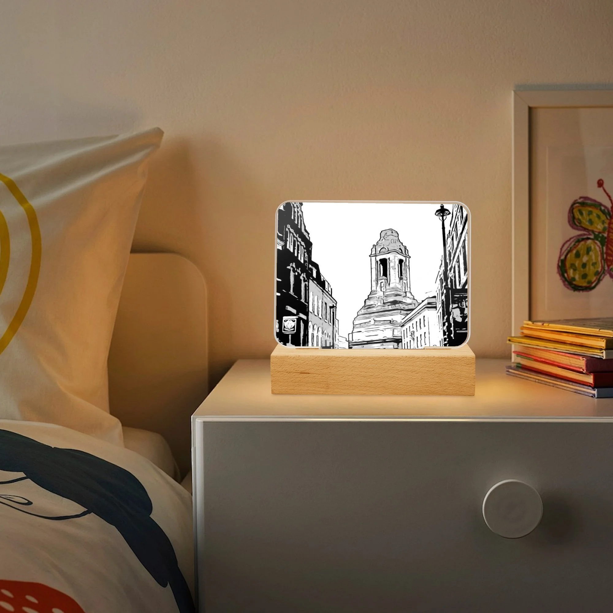 A bedside table with a picture frame displaying a black and white sketch of a city street with a historic monument, next to a bed with a pillow and colorful bedding.