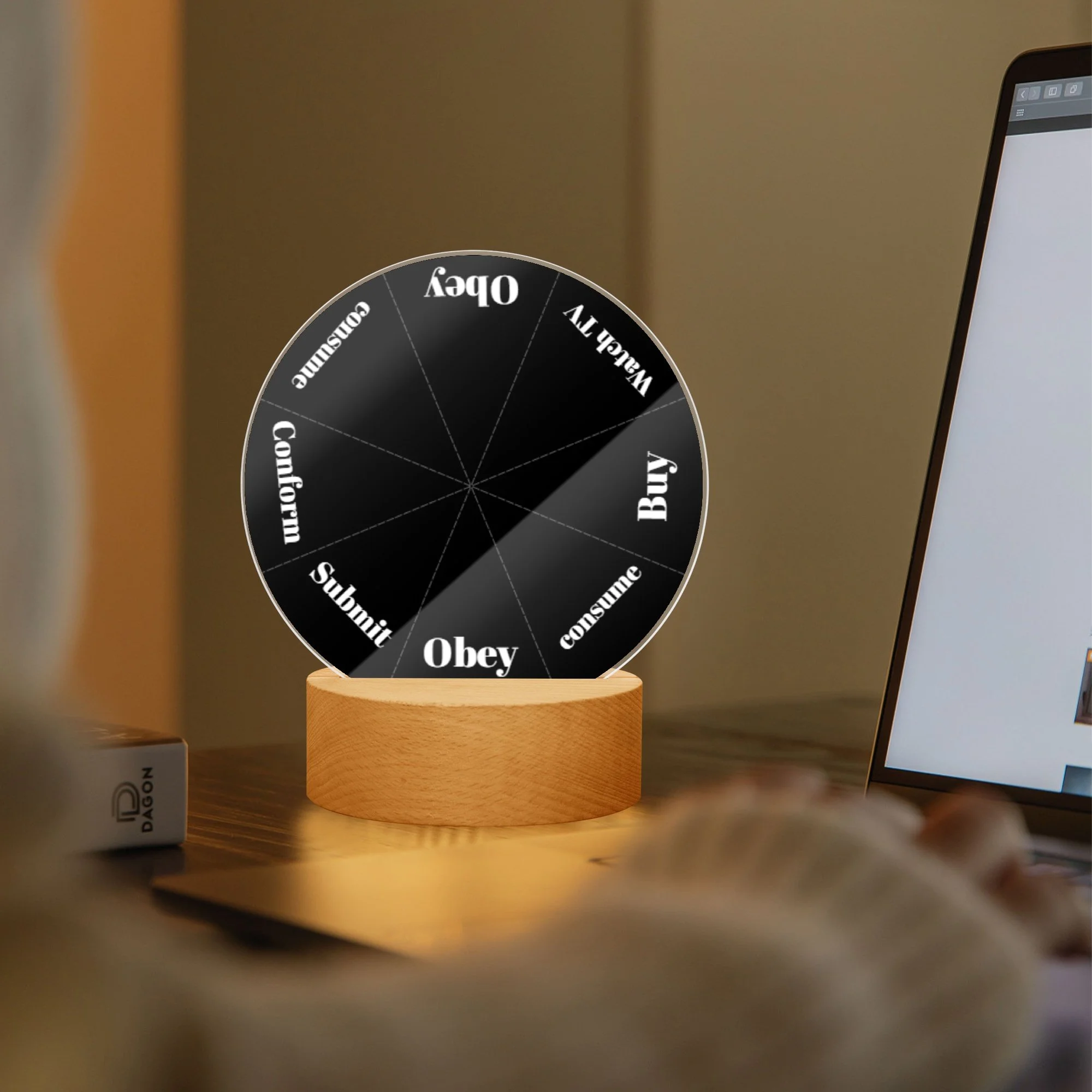 A digital roulette wheel with options like buy, obey, submii, conform, obey, wallify, buy, and debate, displayed on a desk near a laptop