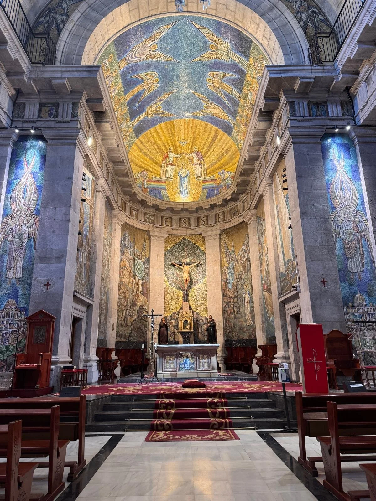 Interior de una iglesia con un altar decorado, crucifijo y mosaicos religiosos en las paredes y techo.