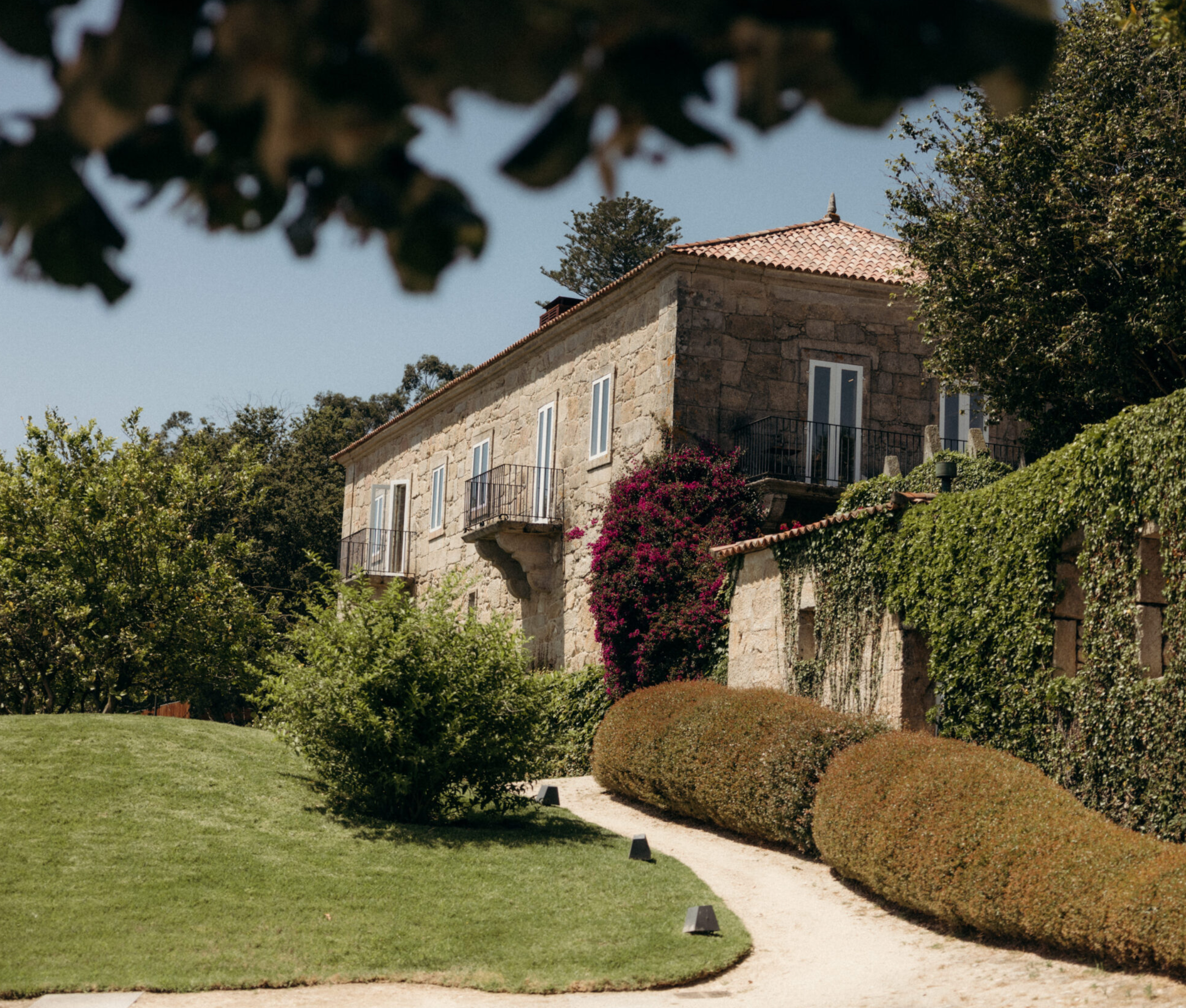 Casa de piedra con balcones, rodeada de un jardín con arbustos y árboles, con un camino de grava