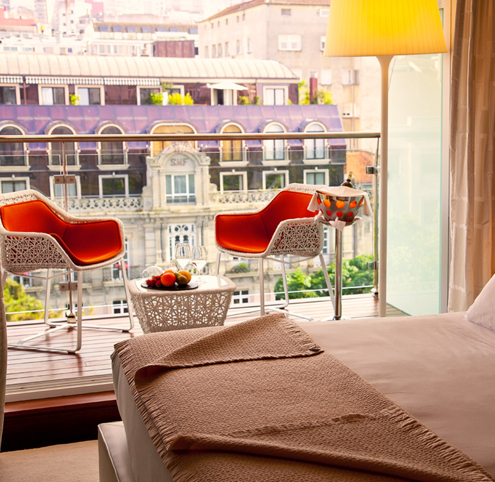 Vista desde una habitación de hotel hacia un balcón con dos sillones de mimbre con cojines naranjas y una mesa con fruta, en una ciudad con edificios históricos.