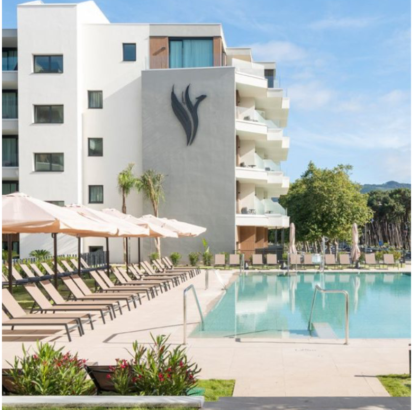Piscina en un complejo hotelero con camastros y sombrillas, edificio moderno con logotipo en la pared, áreas verdes y árboles, cielo despejado.
