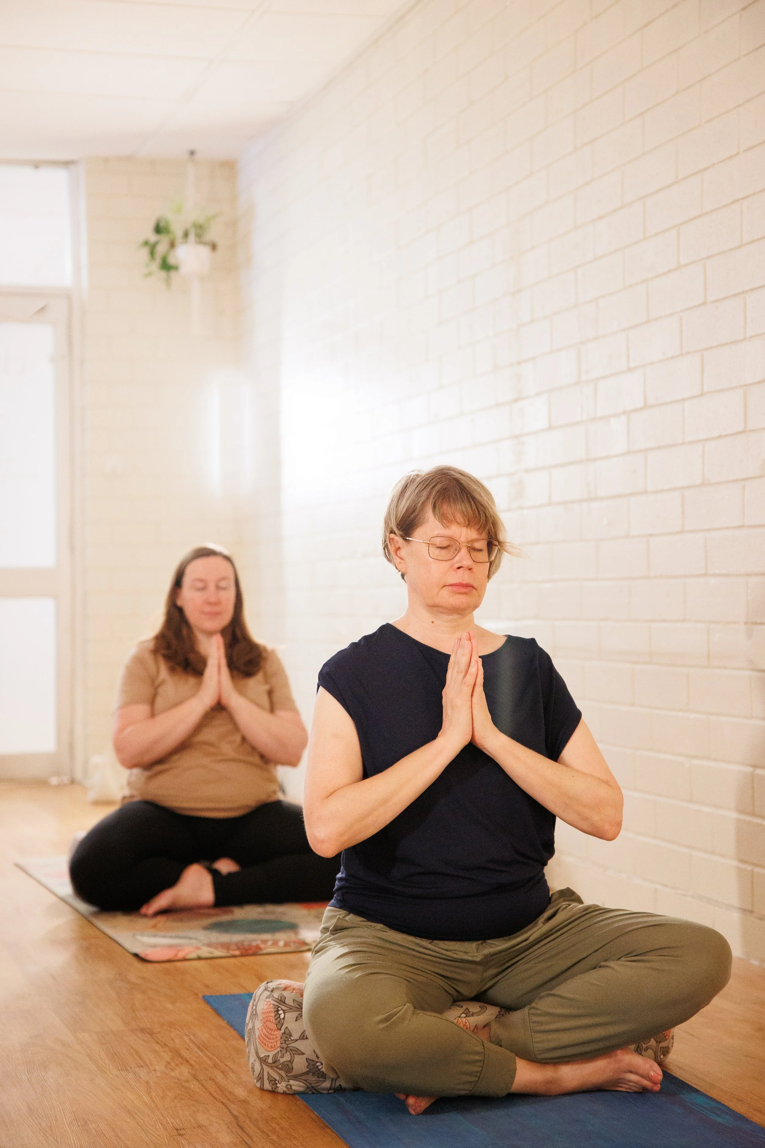 Three women practicing yoga or meditation in a bright room with white brick walls, sitting cross-legged on yoga mats with eyes closed and hands in prayer position.