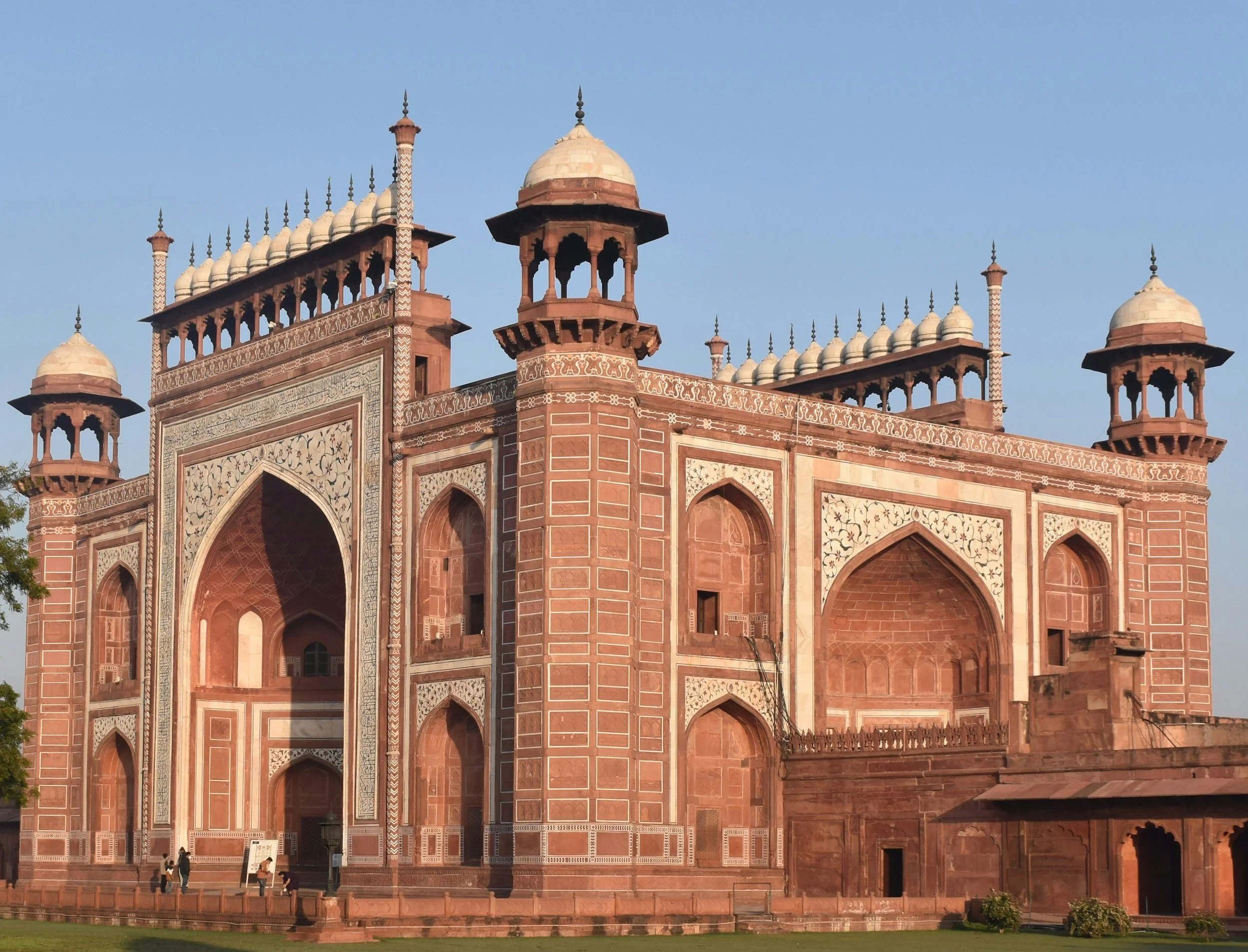 Red sandstone Mughal architecture building with large arched entrance and decorative white inlay work, featuring small domes and minarets under a clear sky.