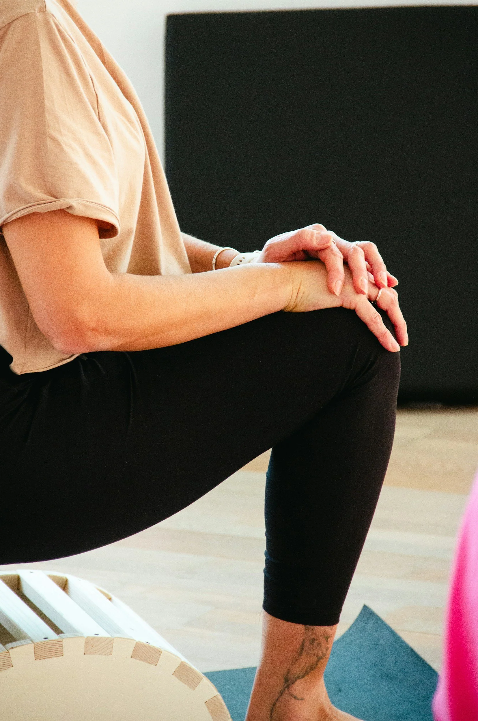 A person wearing a peach-colored t-shirt and black leggings is sitting on the floor, stretching or participating in yoga or exercise. The person's hands are clasped, resting on their knee, and they have a tattoo on their calf. There is a black background and some yoga or exercise equipment nearby.