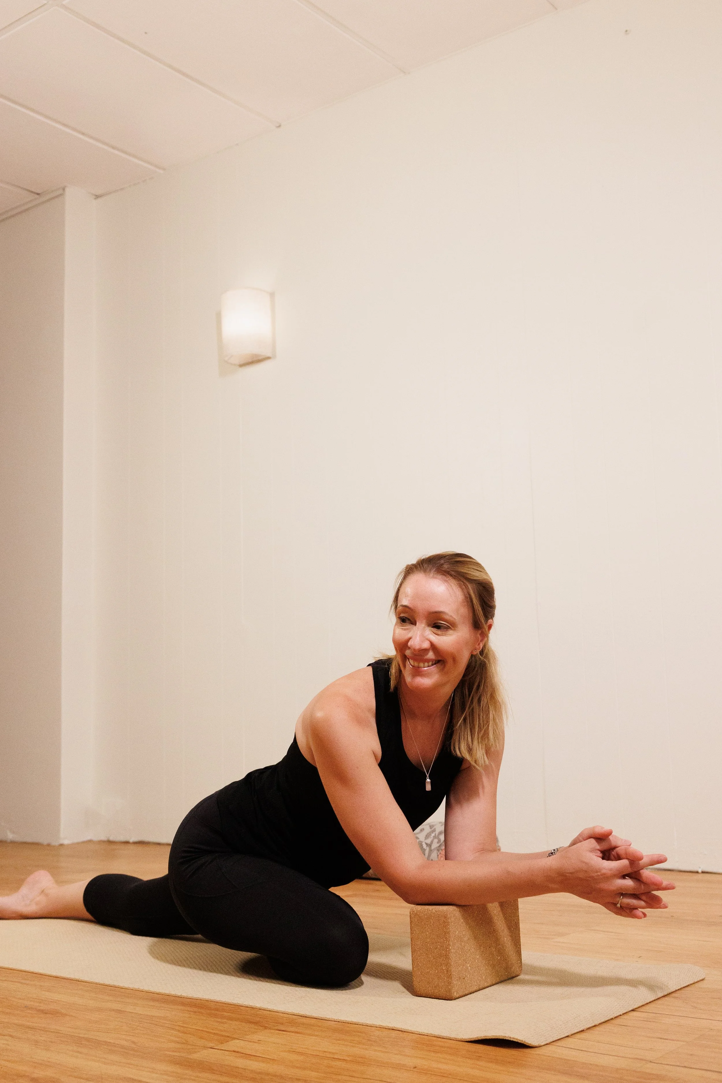 A woman practicing yoga indoors, on a yoga mat, with a cushioned block supporting her arm, smiling and looking to the side.