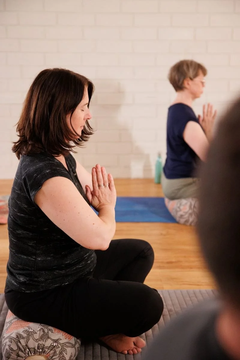 Women practicing meditation or yoga in a seated position with palms pressed together in front of their chest, on yoga mats in a room with a white brick wall.