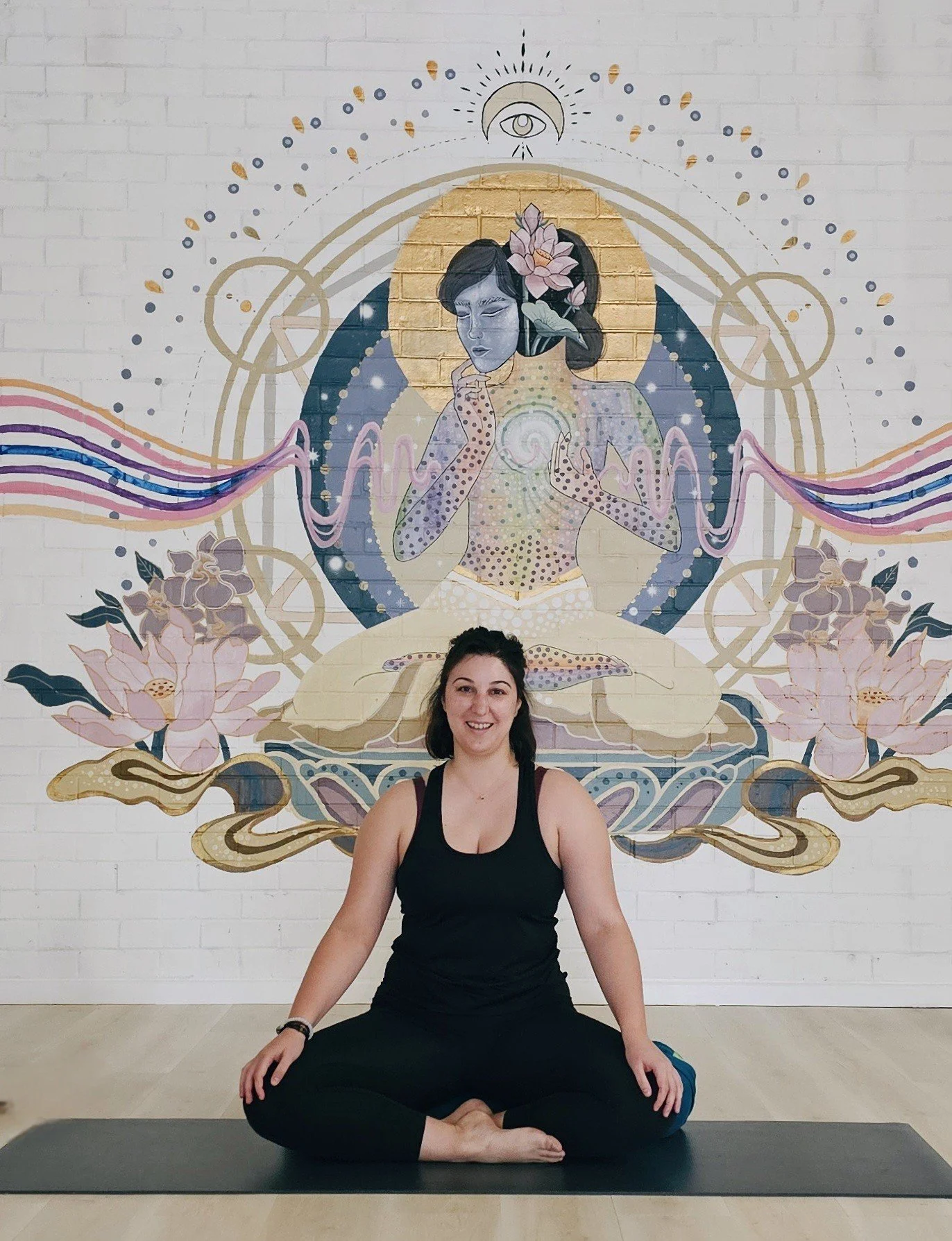 A woman sitting cross-legged on a yoga mat in front of a mural of a meditating figure with a lotus flower in her hair, surrounded by celestial symbols and flowers.